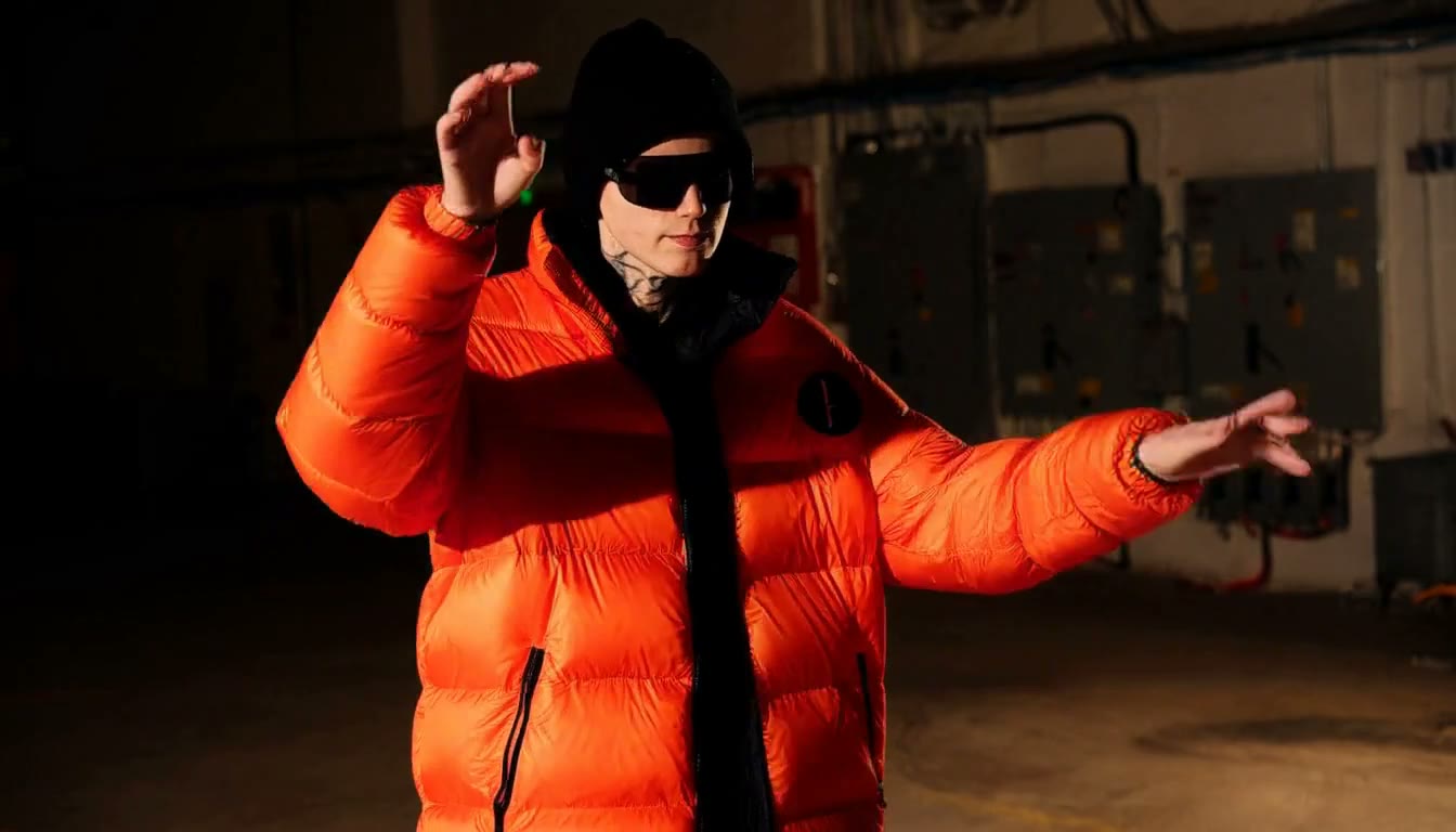 MiniMax H3 AI video example showing a person in an orange puffer jacket and sunglasses is making expressive hand gestures in a dimly lit industrial setting